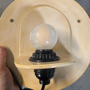 Pu&ograve; includere: Una lampada da parete con una piastra posteriore in ceramica circolare di colore giallo pallido con due fori di montaggio. Un portalampada nero sostiene una lampadina rotonda in vetro smerigliato, posizionata su un piccolo ripiano in ceramica.