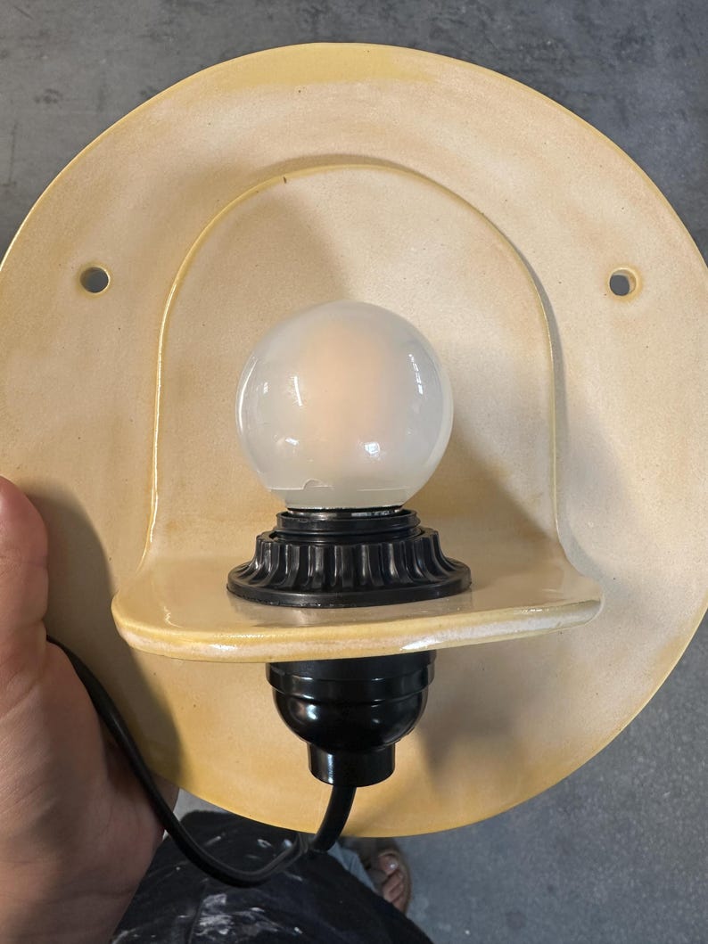 Pu&ograve; includere: Una lampada da parete con una piastra posteriore in ceramica circolare di colore giallo pallido con due fori di montaggio. Un portalampada nero sostiene una lampadina rotonda in vetro smerigliato, posizionata su un piccolo ripiano in ceramica.