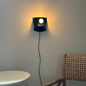 May include: A black wall-mounted sconce featuring a curved, shield-like metal backplate and a single exposed glowing light bulb. A thin black power cord extends downward from the base of the fixture, which is mounted on a plain light-colored wall.