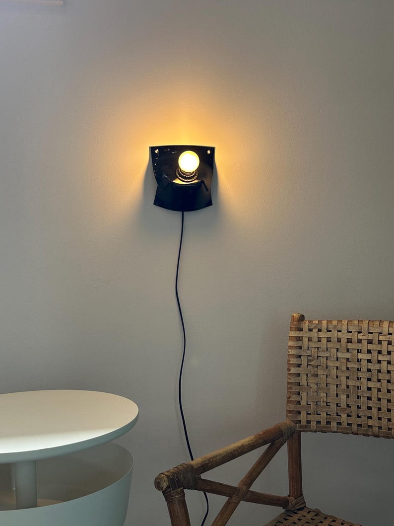 May include: A black wall-mounted sconce featuring a curved, shield-like metal backplate and a single exposed glowing light bulb. A thin black power cord extends downward from the base of the fixture, which is mounted on a plain light-colored wall.