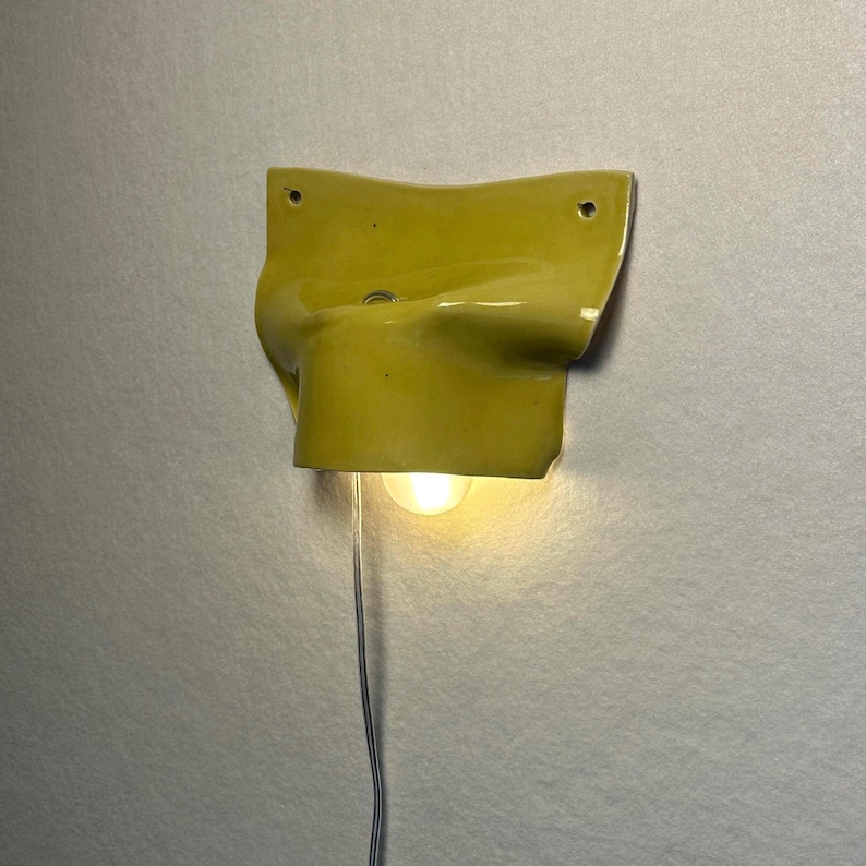 May include: A yellow ceramic wall sconce with a bulb. The sconce has a unique, curved design and is mounted on a light gray wall. The bulb is lit, and a wire hangs down.