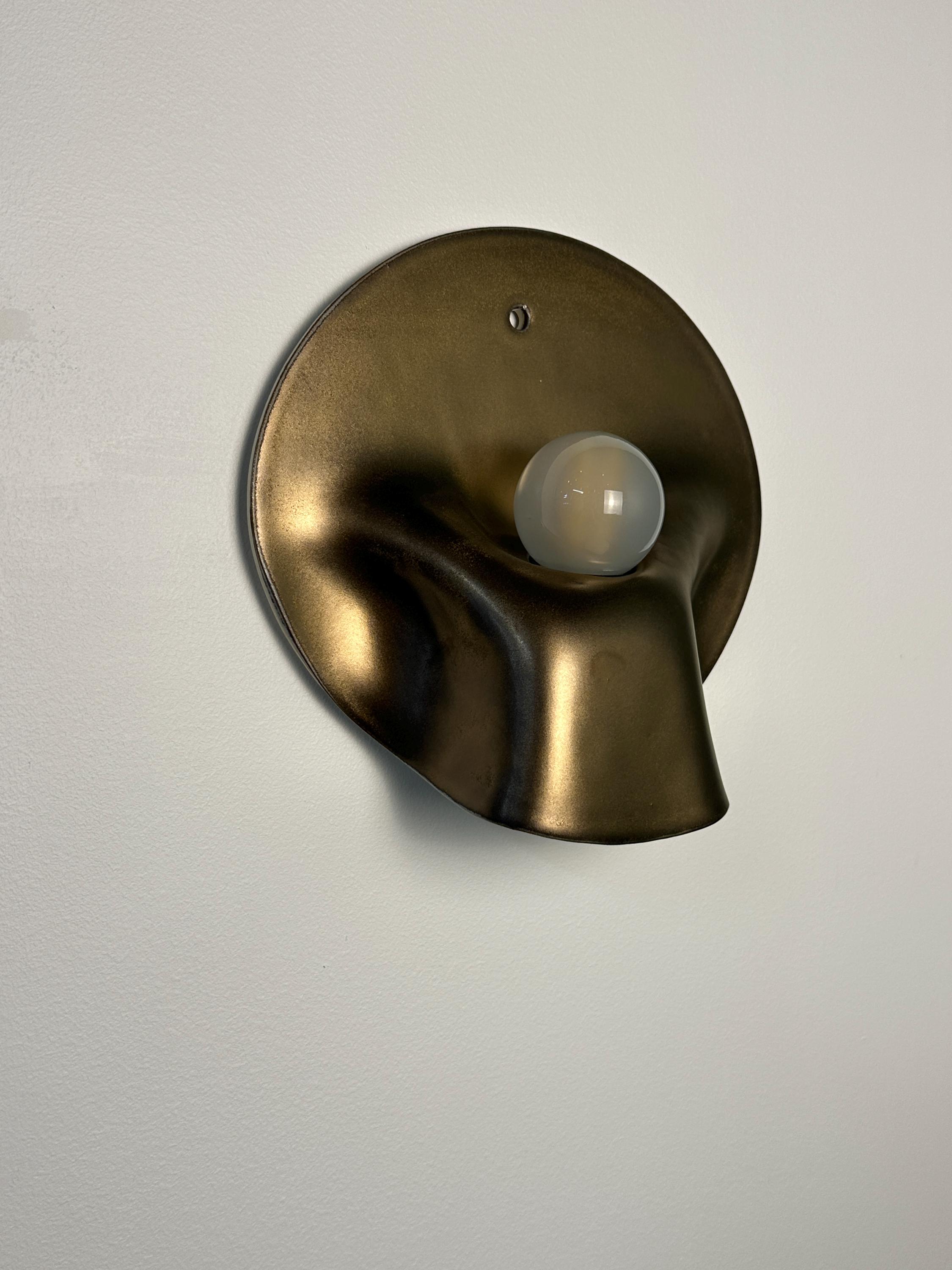 May include: A wall-mounted light fixture featuring a circular, bronze-toned metallic backplate with a fluid, organic, and undulating shape. A single, spherical, frosted white glass light bulb is positioned in the center of the curved metal form.