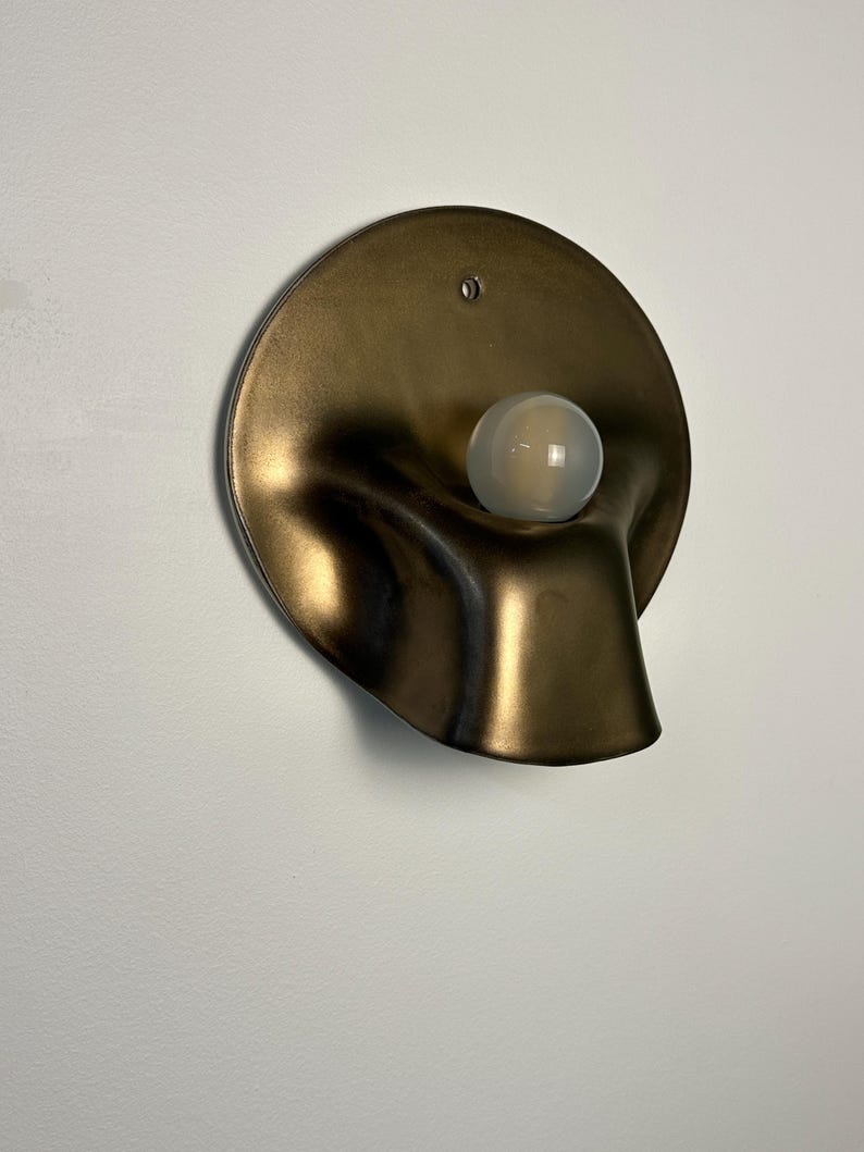 May include: A wall-mounted light fixture featuring a circular, bronze-toned metallic backplate with a fluid, organic, and undulating shape. A single, spherical, frosted white glass light bulb is positioned in the center of the curved metal form.