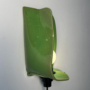 Pu&ograve; includere: Una lampada da parete con un paralume in ceramica verde brillante dalla forma organica curva che ricorda una foglia. L'apparecchio include un portalampada nero e una lampadina bianca posizionata all'interno, collegata da un cavo nero.