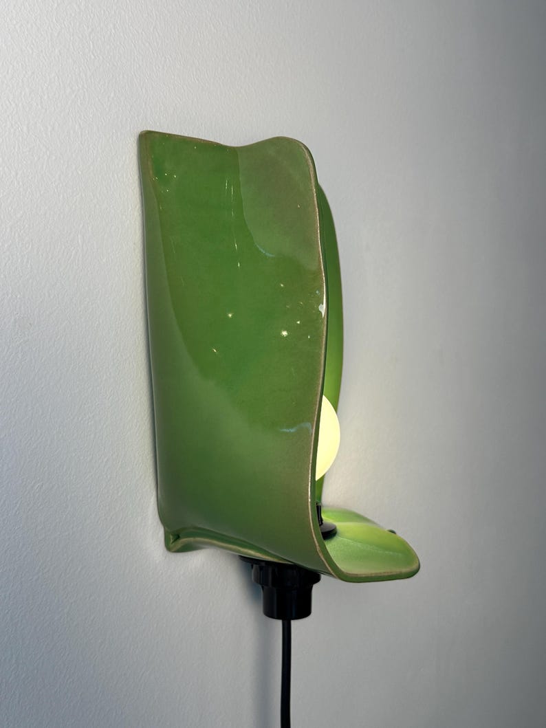 Pu&ograve; includere: Una lampada da parete con un paralume in ceramica verde brillante dalla forma organica curva che ricorda una foglia. L'apparecchio include un portalampada nero e una lampadina bianca posizionata all'interno, collegata da un cavo nero.