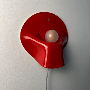May include: A wall-mounted lamp featuring a glossy, vibrant red, organic-shaped shade with a smooth, undulating surface. A single, spherical, frosted white light bulb is positioned within the center of the red form. A thin, clear power cord extends downward.