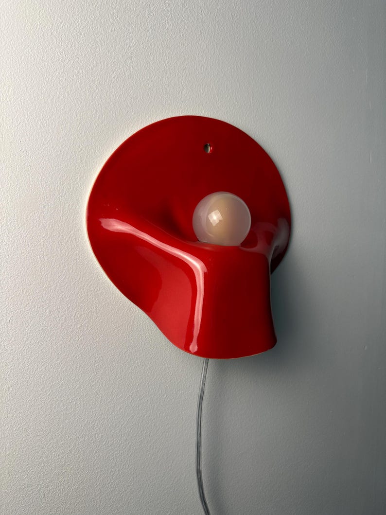 May include: A wall-mounted lamp featuring a glossy, vibrant red, organic-shaped shade with a smooth, undulating surface. A single, spherical, frosted white light bulb is positioned within the center of the red form. A thin, clear power cord extends downward.