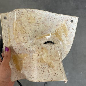 May include: A curved, off-white wall sconce featuring a speckled brown pattern throughout its surface. The ceramic-like fixture has two circular mounting holes at the top corners and a central opening for a black electrical cord and plug.