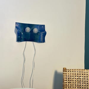 May include: A wall-mounted light fixture featuring a rectangular, wavy blue glass panel with two circular white light bulbs attached to the front. Two thin, transparent electrical cords hang vertically from the bottom of the blue glass panel.