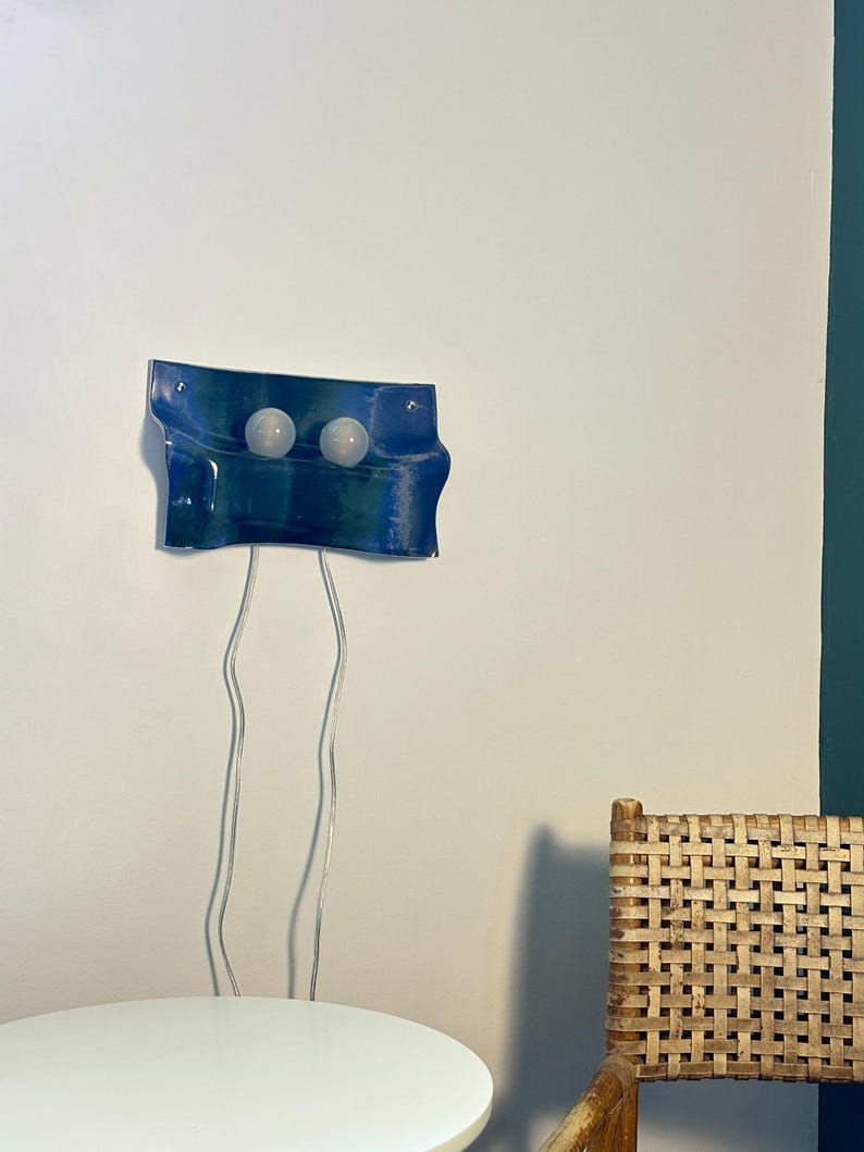 May include: A wall-mounted light fixture featuring a rectangular, wavy blue glass panel with two circular white light bulbs attached to the front. Two thin, transparent electrical cords hang vertically from the bottom of the blue glass panel.