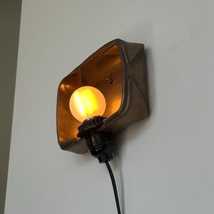 May include: A wall-mounted light fixture with a warm, glowing bulb. The fixture has a bronze-colored, geometric shade and a black base. The light is plugged into a wall outlet with a black cord.