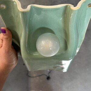 May include: A wall-mounted light fitting featuring a sage green ceramic base with a wavy, organic shape and two circular mounting holes at the top corners. A single, round, frosted white glass light bulb is centred on the ceramic surface.