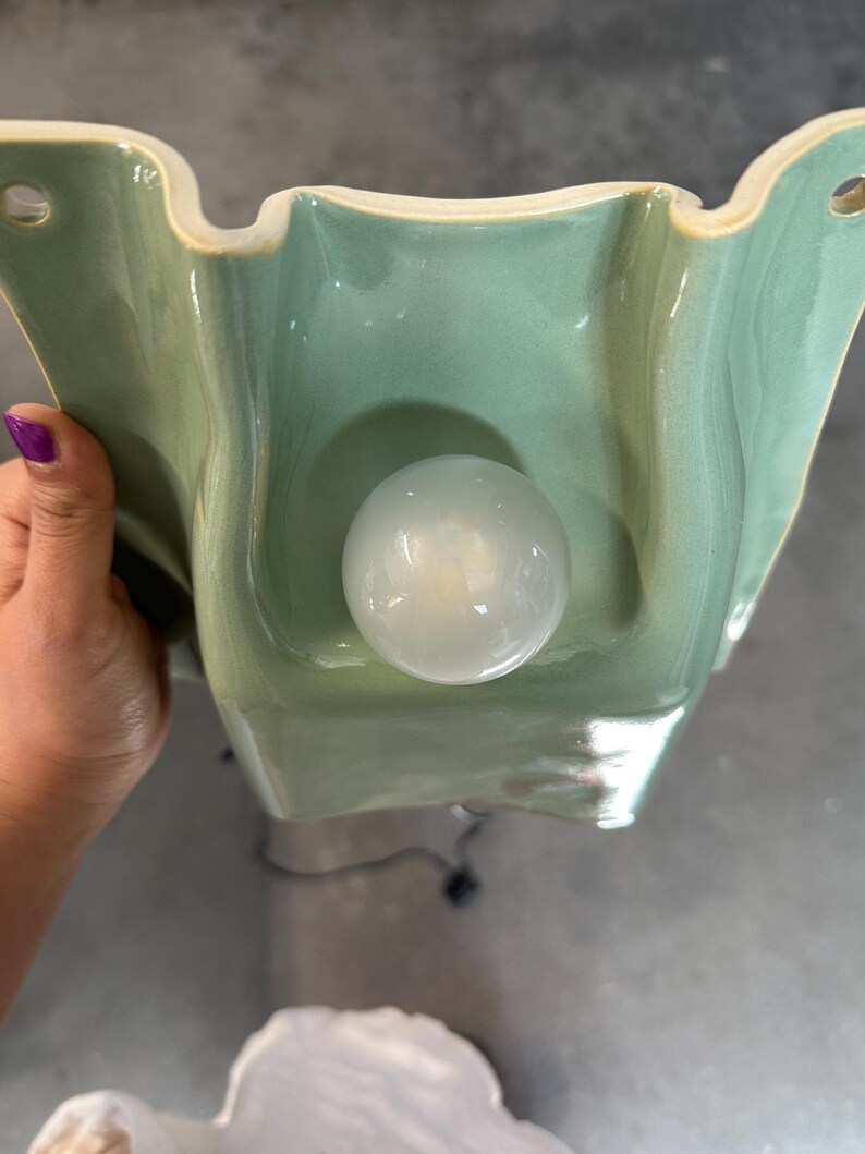 May include: A wall-mounted light fitting featuring a sage green ceramic base with a wavy, organic shape and two circular mounting holes at the top corners. A single, round, frosted white glass light bulb is centred on the ceramic surface.