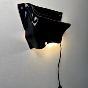 Puede incluir: Una l&aacute;mpara de pared con una forma negra brillante, ondulada y similar a una tela. El accesorio presenta una superficie lisa y reflectante con curvas suaves y emite una luz c&aacute;lida y brillante desde su parte inferior. Un cable negro cuelga.