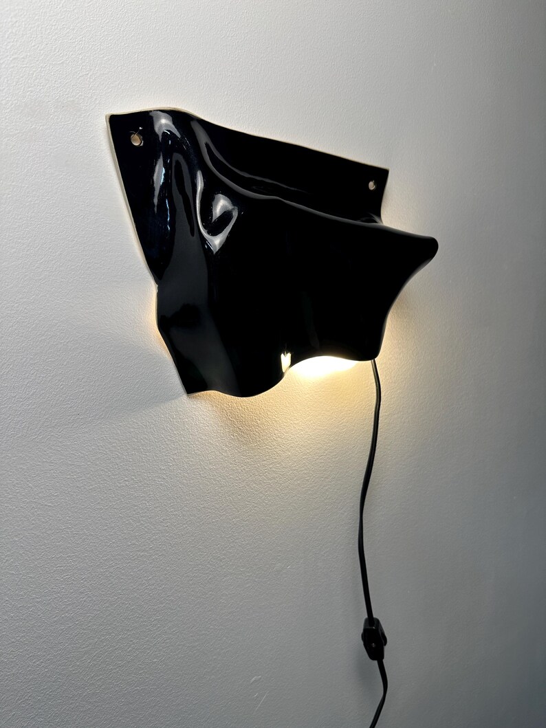 Puede incluir: Una l&aacute;mpara de pared con una forma negra brillante, ondulada y similar a una tela. El accesorio presenta una superficie lisa y reflectante con curvas suaves y emite una luz c&aacute;lida y brillante desde su parte inferior. Un cable negro cuelga.