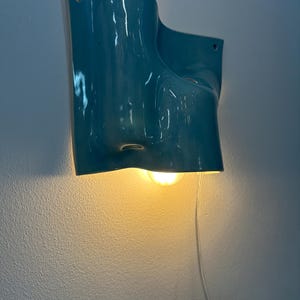 May include: A wall-mounted light fitting with a glossy teal ceramic shade featuring an organic, undulating form. The fitting includes a single warm-toned light bulb emitting a soft glow from beneath the curved structure and a clear power cord.