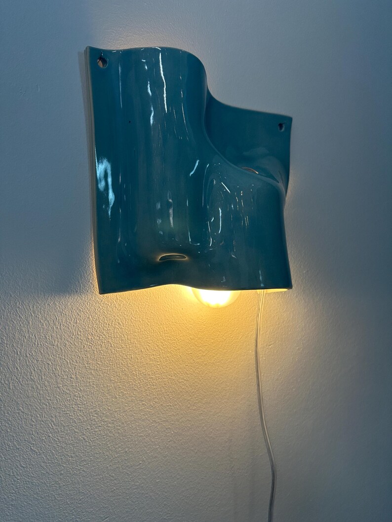 May include: A wall-mounted light fitting with a glossy teal ceramic shade featuring an organic, undulating form. The fitting includes a single warm-toned light bulb emitting a soft glow from beneath the curved structure and a clear power cord.