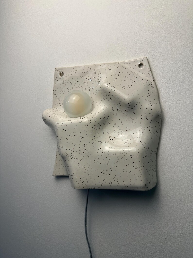 May include: A wall-mounted lamp featuring a white ceramic base with a black speckled pattern. The base has an irregular, molded shape with a circular cutout holding a frosted glass globe light bulb. A thin black power cord extends from the bottom.