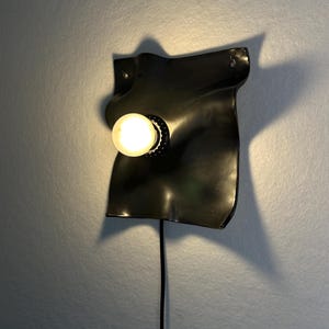 May include: A wall-mounted lamp featuring a glossy black, irregularly shaped, undulating ceramic or metal plate. A single round, glowing light bulb is centered on the surface, and a black power cord extends downward from the base of the fixture.
