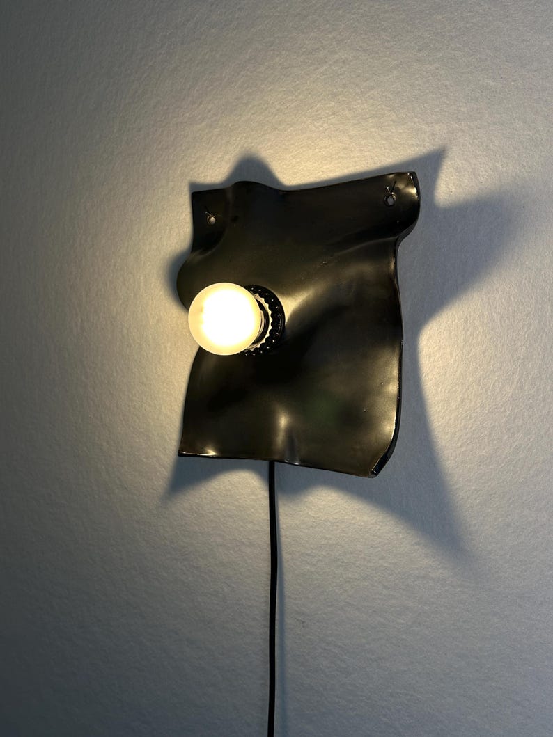 May include: A wall-mounted lamp featuring a glossy black, irregularly shaped, undulating ceramic or metal plate. A single round, glowing light bulb is centered on the surface, and a black power cord extends downward from the base of the fixture.