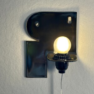 May include: A wall-mounted lamp featuring a glossy black ceramic base shaped like the letter R. The fixture includes a small, round, frosted white light bulb set into a black socket, with a thin, clear power cord extending downwards from the base.