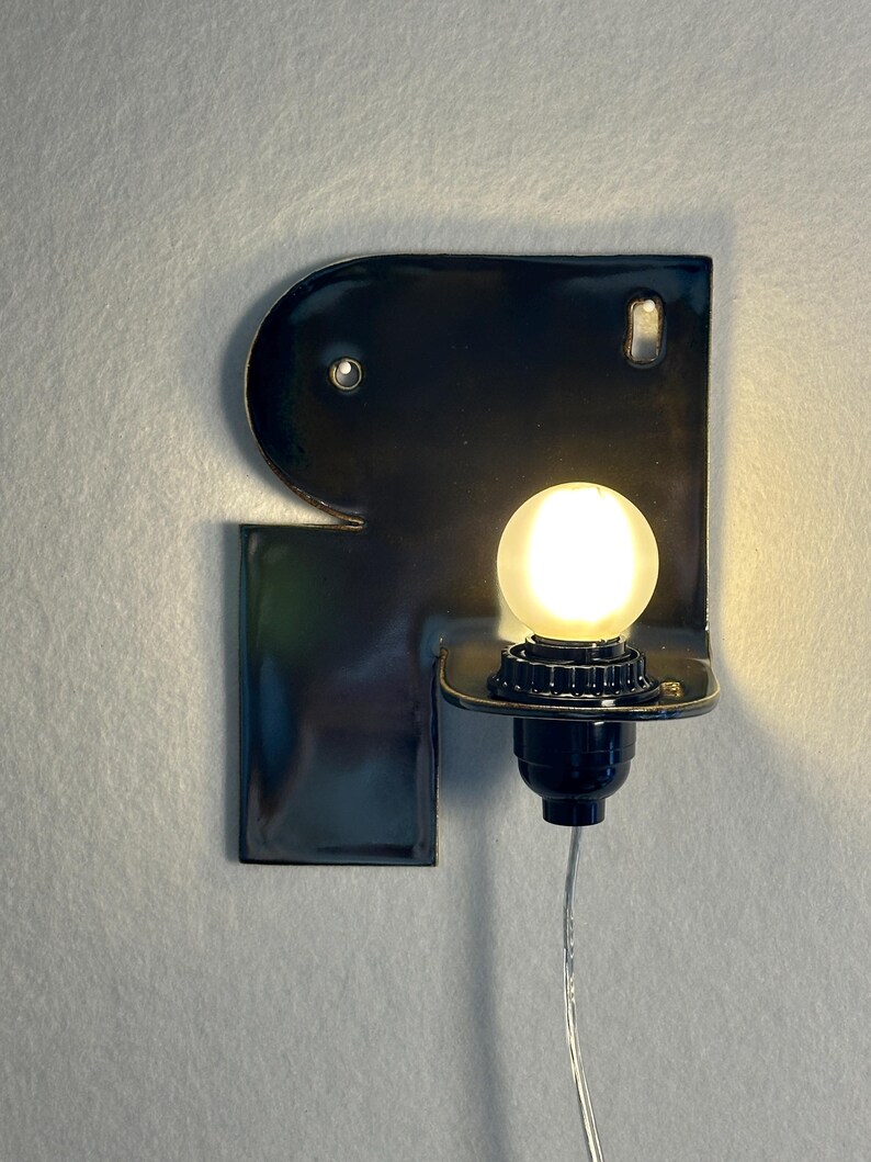 May include: A wall-mounted lamp featuring a glossy black ceramic base shaped like the letter R. The fixture includes a small, round, frosted white light bulb set into a black socket, with a thin, clear power cord extending downwards from the base.