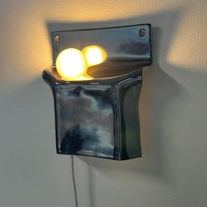 Peut inclure: Une applique murale en c&eacute;ramique avec une finition brillante, sombre, bleu et noir marbr&eacute;. Le luminaire pr&eacute;sente une forme incurv&eacute;e en poche qui contient une ampoule sph&eacute;rique unique et lumineuse, projetant une lumi&egrave;re chaude et vive.