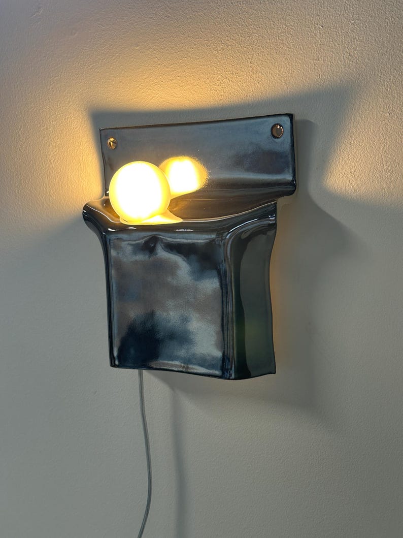 Peut inclure: Une applique murale en c&eacute;ramique avec une finition brillante, sombre, bleu et noir marbr&eacute;. Le luminaire pr&eacute;sente une forme incurv&eacute;e en poche qui contient une ampoule sph&eacute;rique unique et lumineuse, projetant une lumi&egrave;re chaude et vive.