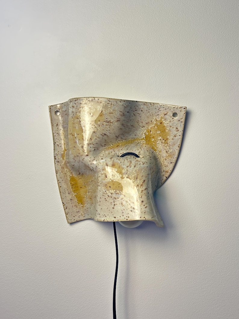 May include: A wall-mounted ceramic light fixture with an irregular, folded shape. The surface features a speckled cream and beige finish with prominent yellow glazed accents. A black power cord extends from the bottom, and two mounting holes are visible.