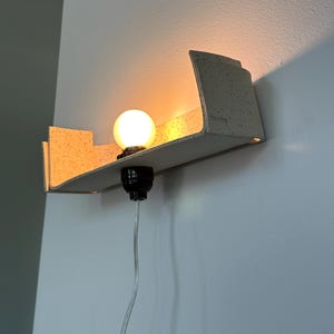 May include: A modern wall-mounted light fixture with a speckled, light-colored shelf. A round, illuminated light bulb sits in a black socket. A clear cord hangs down. The fixture is mounted on a white wall.