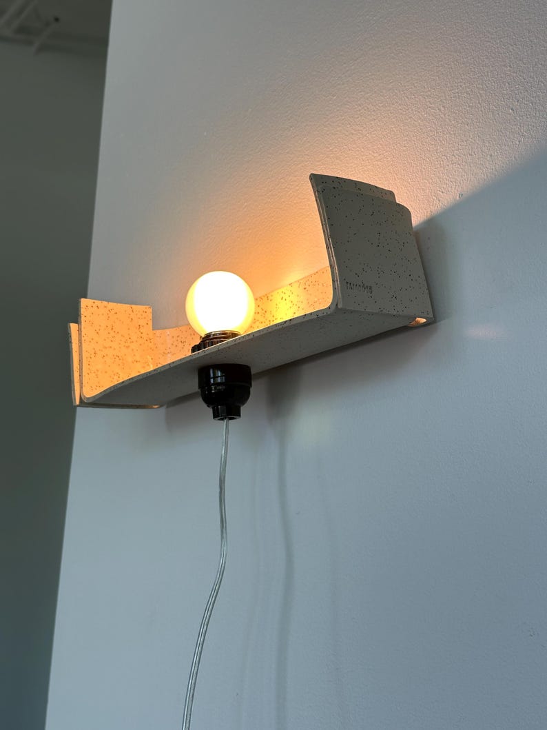 May include: A modern wall-mounted light fixture with a speckled, light-colored shelf. A round, illuminated light bulb sits in a black socket. A clear cord hangs down. The fixture is mounted on a white wall.