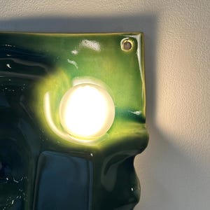 May include: A wall-mounted light fixture featuring a glossy, undulating ceramic panel in shades of deep forest green and lime green. A single, spherical, frosted white glass bulb is centered on the panel, emitting a warm, bright glow.