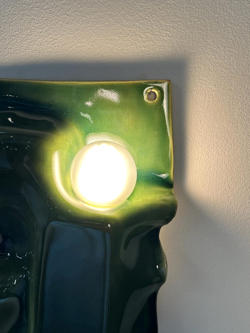 May include: A wall-mounted light fixture featuring a glossy, undulating ceramic panel in shades of deep forest green and lime green. A single, spherical, frosted white glass bulb is centered on the panel, emitting a warm, bright glow.
