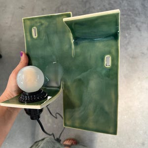 May include: A wall-mounted ceramic lamp featuring a glossy, variegated green glaze. The structure consists of two rectangular, offset panels with two rectangular cutouts. A black socket holds a round, frosted white light bulb on the lower shelf.