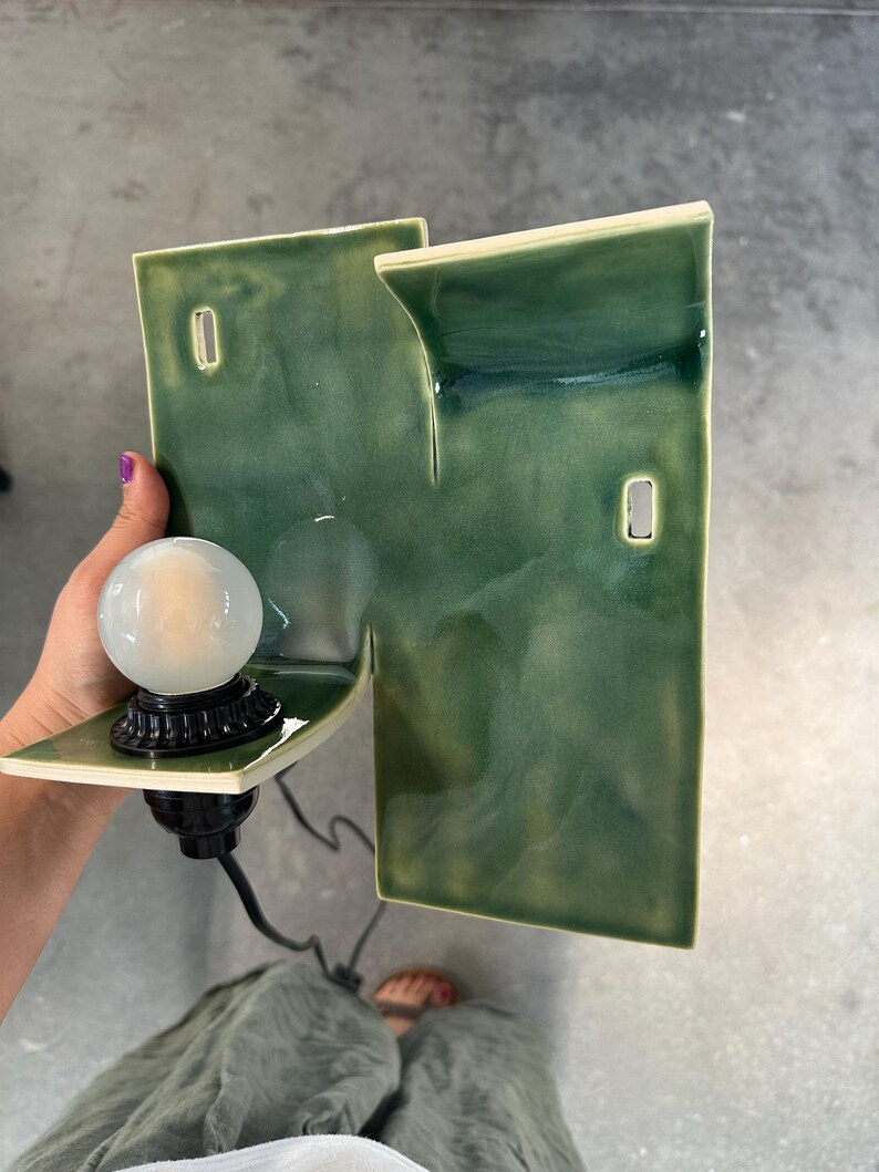 May include: A wall-mounted ceramic lamp featuring a glossy, variegated green glaze. The structure consists of two rectangular, offset panels with two rectangular cutouts. A black socket holds a round, frosted white light bulb on the lower shelf.