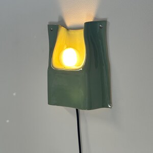 May include: A wall-mounted ceramic sconce featuring a glossy sage green exterior and a smooth, cream-coloured interior basin. The fixture houses a single glowing light bulb that casts a warm upward beam, and it includes a black power cord.