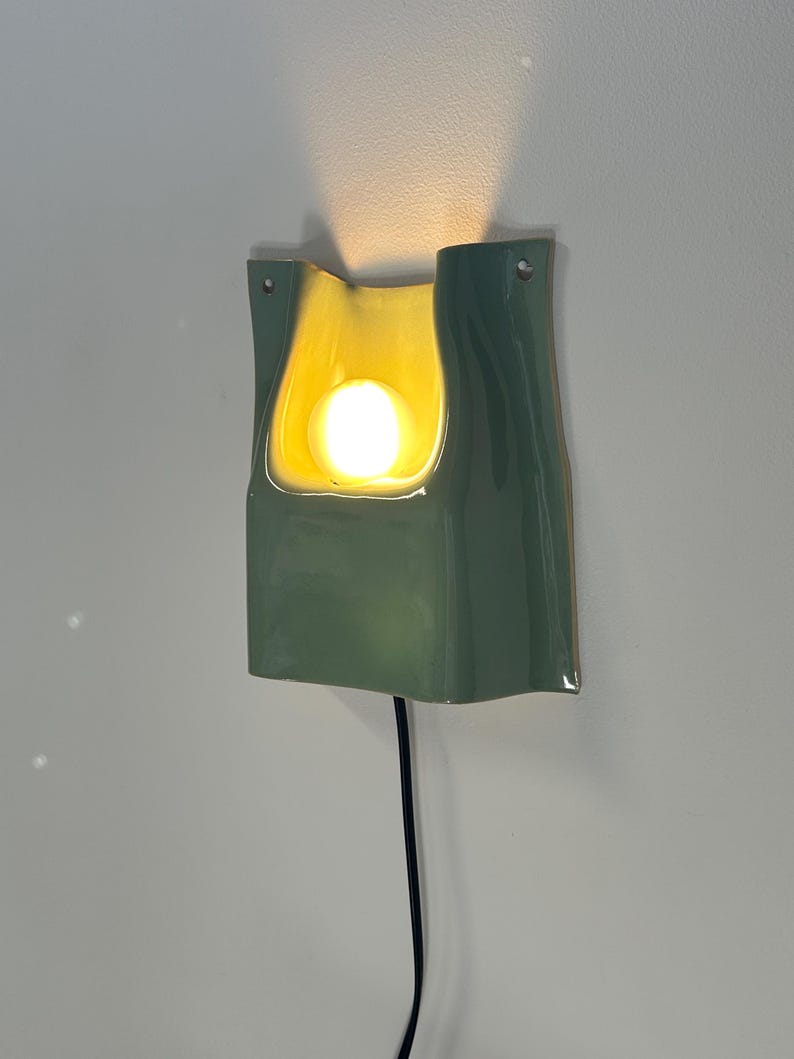 May include: A wall-mounted ceramic sconce featuring a glossy sage green exterior and a smooth, cream-coloured interior basin. The fixture houses a single glowing light bulb that casts a warm upward beam, and it includes a black power cord.