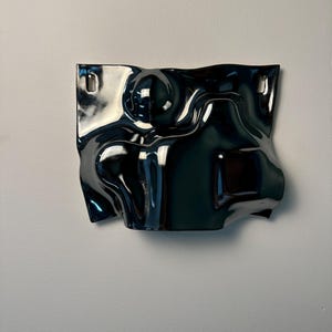 Pu&ograve; includere: Un pezzo scultoreo da parete con una finitura nera lucida e riflettente. L'oggetto presenta una superficie irregolare e ondulata con curve e rientranze organiche. Due aperture rettangolari sono posizionate vicino agli angoli superiori.