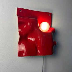 May include: A wall-mounted lamp featuring a glossy, bright red, undulating square panel with a smooth, organic, wave-like surface. A single, round, glowing white light bulb is embedded within a circular cutout on the right side of the red panel.
