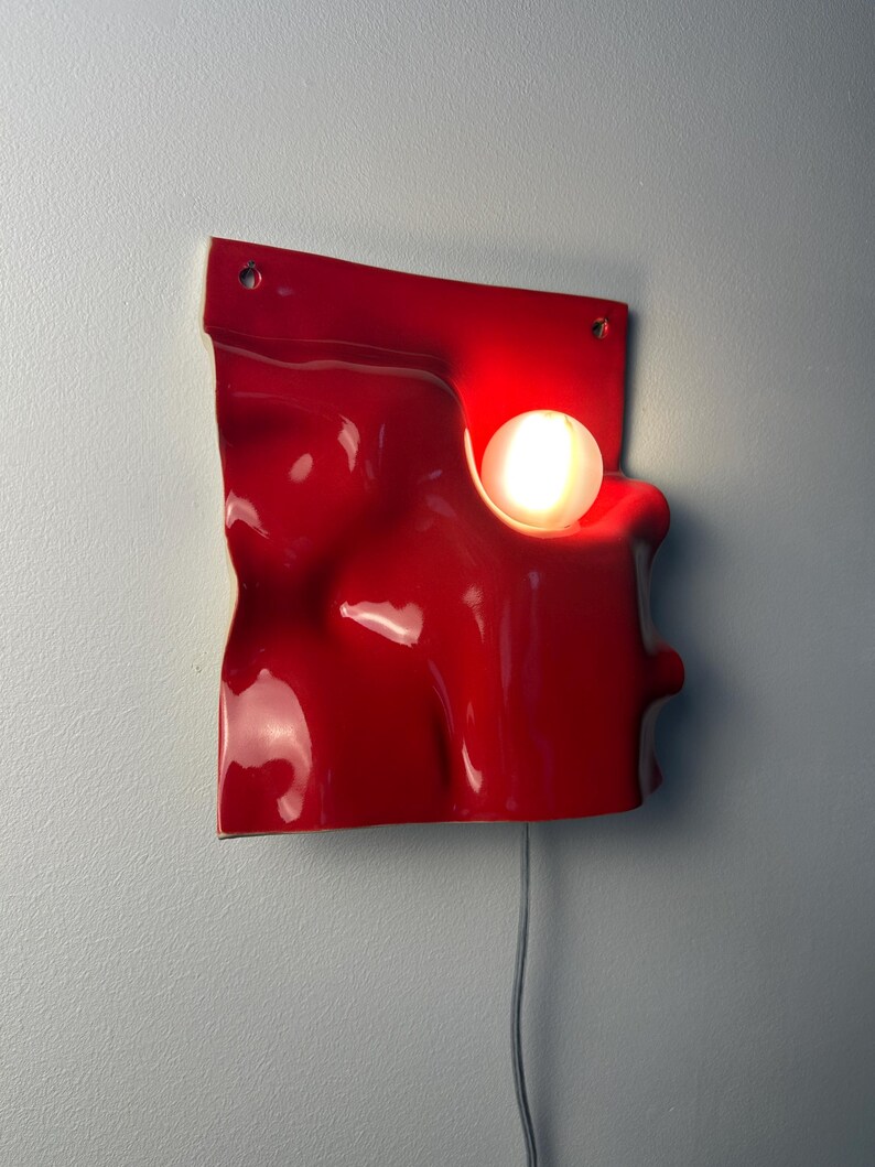 May include: A wall-mounted lamp featuring a glossy, bright red, undulating square panel with a smooth, organic, wave-like surface. A single, round, glowing white light bulb is embedded within a circular cutout on the right side of the red panel.