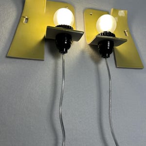 May include: A pair of wall-mounted lamps featuring pale yellow, curved metal backplates. Each lamp has a black socket holding a round, glowing white light bulb. A clear electrical cord extends downward from the base of each black light socket.
