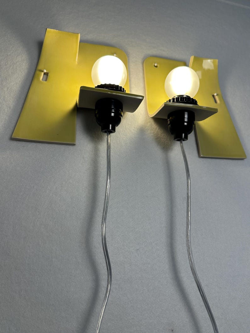May include: A pair of wall-mounted lamps featuring pale yellow, curved metal backplates. Each lamp has a black socket holding a round, glowing white light bulb. A clear electrical cord extends downward from the base of each black light socket.