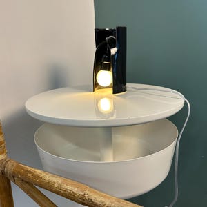 May include: A white tiered side table with a circular top and a deep bowl-shaped lower shelf, featuring a black ceramic sculptural lamp with an exposed glowing light bulb placed on the top surface, connected by a thin white electrical cord.
