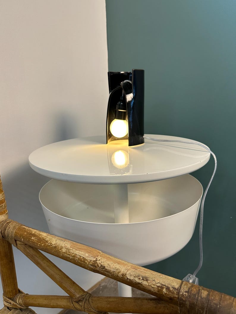May include: A white tiered side table with a circular top and a deep bowl-shaped lower shelf, featuring a black ceramic sculptural lamp with an exposed glowing light bulb placed on the top surface, connected by a thin white electrical cord.