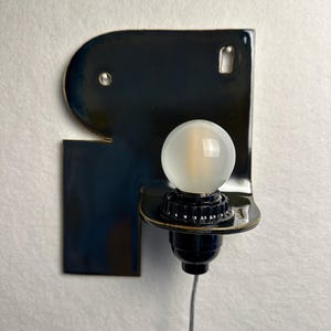 May include: A wall-mounted lamp featuring a glossy, dark navy blue ceramic backplate with an irregular, rounded shape. A black socket holds a round, frosted white light bulb, and a thin, clear electrical cord extends downwards from the base.