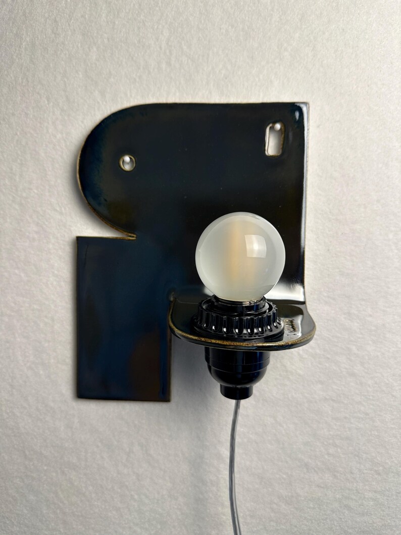 May include: A wall-mounted lamp featuring a glossy, dark navy blue ceramic backplate with an irregular, rounded shape. A black socket holds a round, frosted white light bulb, and a thin, clear electrical cord extends downwards from the base.