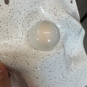 May include: A white, speckled, molded plastic form featuring a smooth, glossy, spherical protrusion in the center. The surface has a textured, uneven, and undulating shape with two small circular holes visible along the top edge of the material.