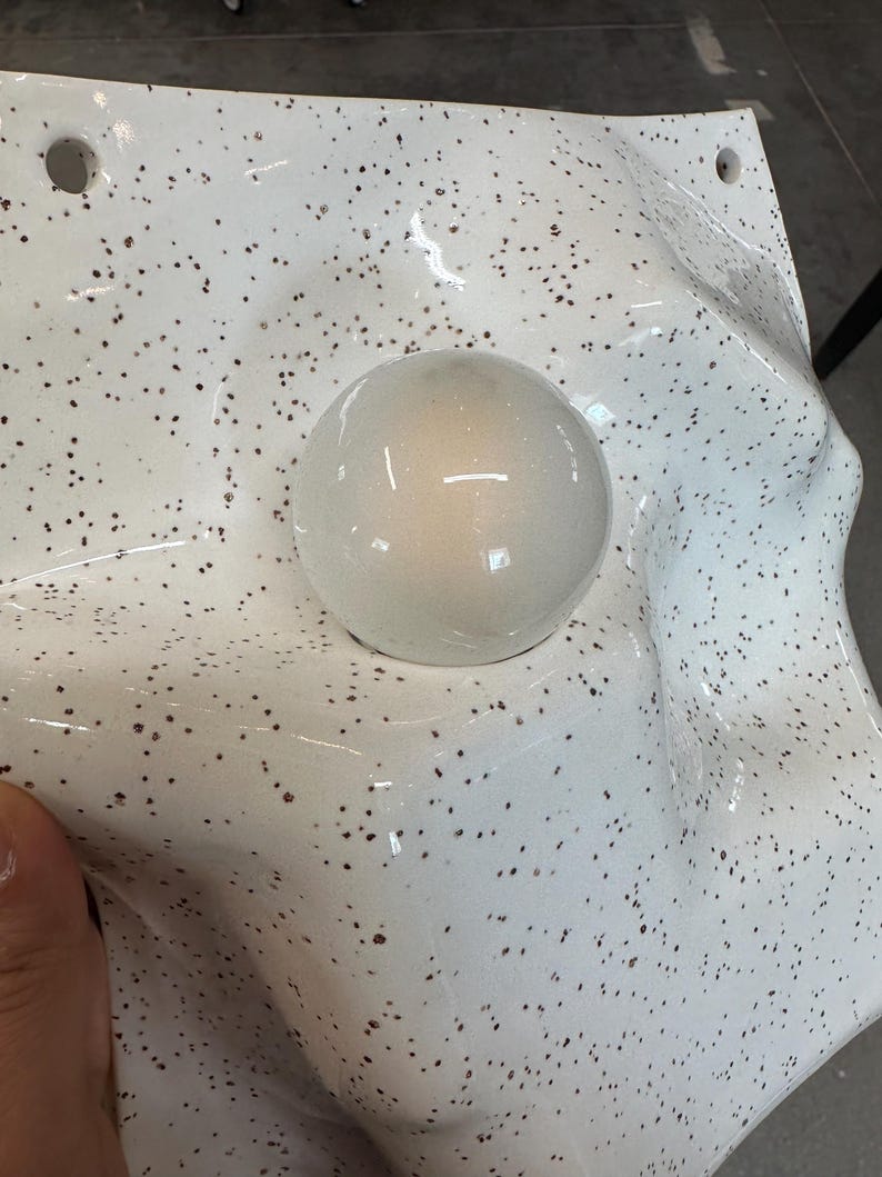 May include: A white, speckled, molded plastic form featuring a smooth, glossy, spherical protrusion in the center. The surface has a textured, uneven, and undulating shape with two small circular holes visible along the top edge of the material.