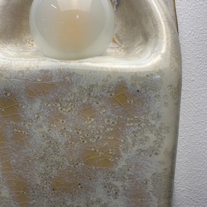 May include: A wall-mounted ceramic light fitting featuring a glossy, cream-coloured glaze with a distinct crystalline, crackled texture in shades of yellow and white. The fitting includes a central, spherical, frosted glass light bulb.