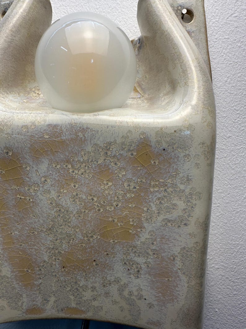 May include: A wall-mounted ceramic light fitting featuring a glossy, cream-coloured glaze with a distinct crystalline, crackled texture in shades of yellow and white. The fitting includes a central, spherical, frosted glass light bulb.